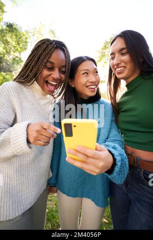 Vertikal. Drei junge Frauen, die telefonieren, sich im Park amüsieren und lachen. Social-Media-App Stockfoto