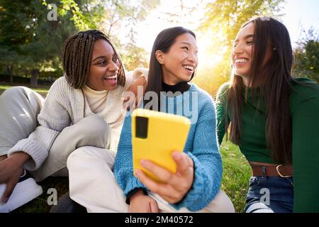 Drei junge Frauen, die ein Handy benutzen, während sie im Park Spaß haben und lachen. Social-Media-App Stockfoto