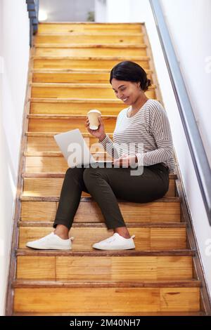 Multitasking gehört zur Arbeit. Foto einer attraktiven jungen Geschäftsfrau, die auf einer hölzernen Treppe sitzt und dabei ihren Laptop benutzt Stockfoto