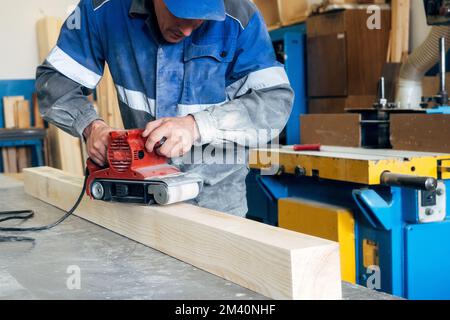 Zimmermann in Arbeitskleidung verarbeitet Holz mit Schleifer. Polieren von Holzoberflächen. Ein Mann mittleren Alters arbeitet auf einer Werkbank in einer Tischlerwerkstatt. Echter Workflow. Stockfoto