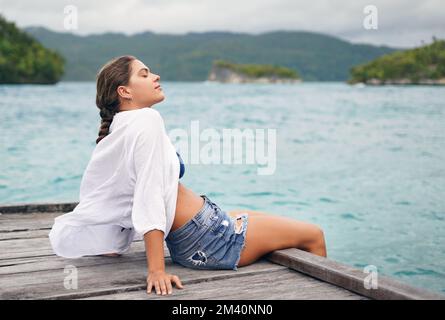 Spüren Sie die Brise. Eine attraktive junge Frau, die am Rande einer Promenade sitzt und während des Urlaubs eine Brise genießt. Stockfoto