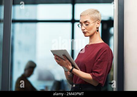 Unternehmens-E-Mails durchgehen. Eine attraktive junge Geschäftsfrau, die eine Brille trägt und ein Tablet benutzt, während sie allein in ihrem Büro steht. Stockfoto