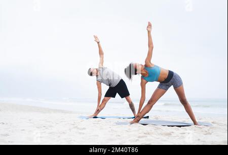 Die frische Luft inspiriert uns, fit zu bleiben. Ein junger Mann und eine junge Frau, die zusammen Yoga am Strand praktizieren. Stockfoto