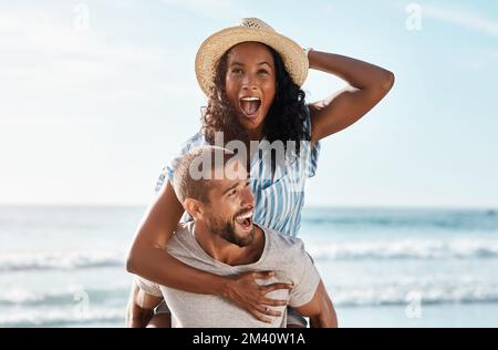 Die Sommer sind so eine glückliche Jahreszeit. Ein junger Mann, der seine Freundin am Strand mit dem Huckepack versorgte. Stockfoto