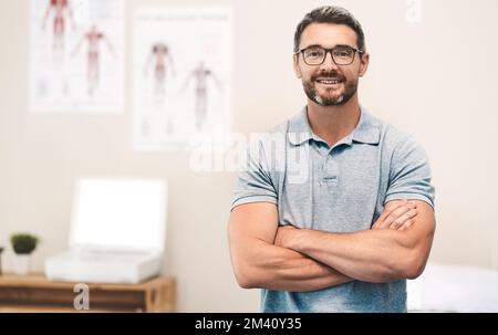 An die Arbeit. Porträt eines gut aussehenden, reifen Physiotherapeuten, der mit gefalteten Armen in seinem Büro posiert. Stockfoto