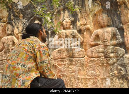 Rückansicht eines langhaarigen jungen Mannes, der vor der Gyalwa Ringna 5 Dhyani Buddha-Felsstatue sitzt, die sich in Zanskar, Ladakh, Indien befindet Stockfoto