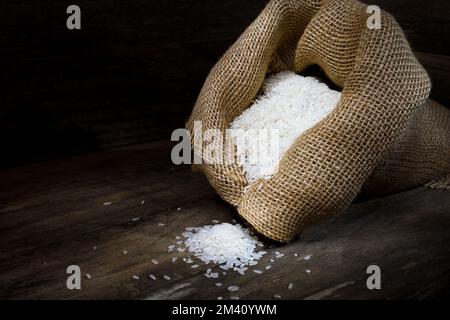 Eine rustikale Szene mit einem hessischen Sack, der mit weißem Langkornreis auf einer hölzernen, kastenähnlichen Umgebung in dunkler Stimmungsbeleuchtung überflutet und in einem Studio aufgenommen wurde Stockfoto