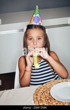 Rate mal, wer heute ein Jahr älter ist. Ein bezauberndes kleines Mädchen, das auf einer Geburtstagsfeier ein Partyhorn bläst. Stockfoto