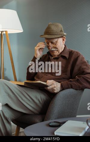 Ein älterer Mann sitzt allein auf einem Stuhl im Wohnzimmer, trägt eine Brille und liest ein Buch. Stockfoto