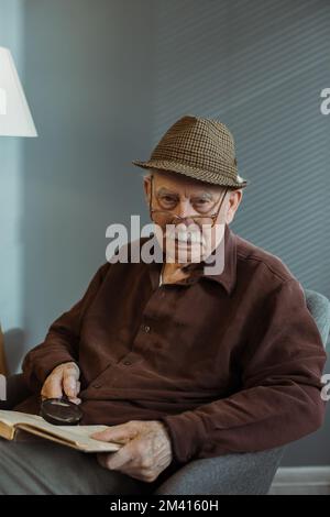 Ein älterer Mann, der allein auf einem Sessel im Wohnzimmer sitzt, eine Brille trägt und ein Buch hält, mit Bedacht in die Kamera schaut Stockfoto