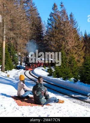 Ein paar Männer und Frauen beobachten die Dampfeisenbahn im Winter im Schnee im Harz-Nationalpark Deutschland, die Dampfeisenbahn Brocken Bahn auf dem Weg durch die Winterlandschaft Stockfoto