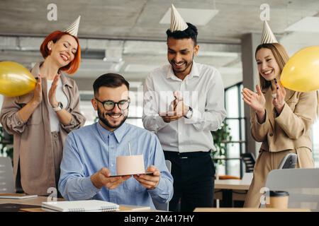 Der junge Mann wird Kerzen auf Kuchen blasen und sich etwas wünschen, während er mit Kollegen Geburtstag feiert. Kollegen, die im Büro Geburtstag feiern Stockfoto