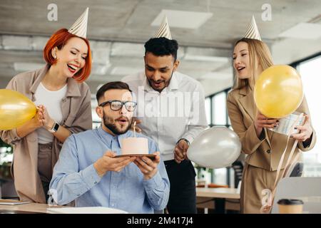 Ein glücklicher Geschäftsmann bläst eine Kerze auf dem Kuchen aus, während er mit weiblichen Kollegen im Büro Geburtstag feiert. Stockfoto