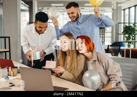 Glückliche Geschäftsfrau, die eine Kerze auf dem Kuchen ausbläst, während sie ihren Geburtstag mit weiblichen Kollegen im Büro feiert. Stockfoto