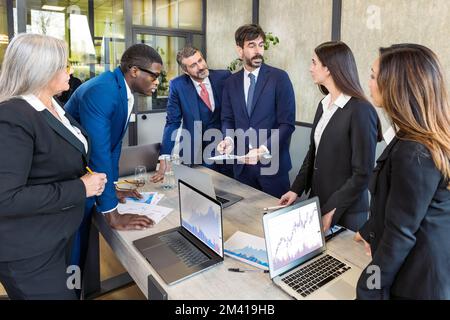 Gruppe von Kollegen aus verschiedenen ethnischen Gruppen in formeller Kleidung, die während des Meetings in einem modernen Büro Diagramme und Planungsarbeiten besprechen und sich am Tisch versammeln Stockfoto