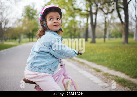 Kinder, Fahrrad fahren und lernen, mit einem Mädchen im Park auf ihrem Fahrrad zu fahren, während sie einen Helm tragen, um draußen sicher zu sein. Sommer, Radfahren und Kinder mit einem Stockfoto