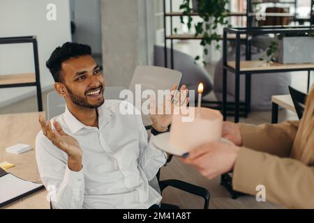 Ein junger Hindu-Mann ist dabei, Kerzen auf einem Kuchen auszublasen und sich während einer Geburtstagsfeier mit Kollegen etwas zu wünschen. Kollegen feiern eine Geburt Stockfoto