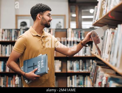 Universität, Student oder Mann mit bibel in einer Bibliothek, um spirituelle Religionskenntnisse, Gebete oder Christus Bücherregalsuche zu studieren. Lächeln, Schule oder glücklich Stockfoto