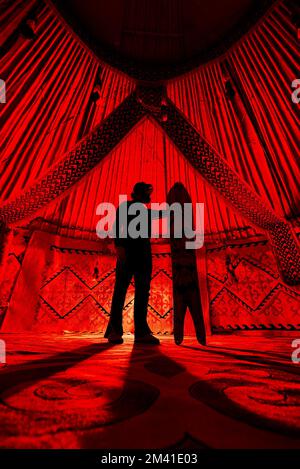 Silhouette von man Holding mit geteiltem Snowboard im Nomadischen Haus von Yurt gegen Teppich mit ethnischen Mustern, beleuchtet von rotem Innenraum. Backcountry ri Stockfoto