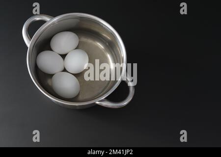 Gekochte Hühnereier in einem Topf. Metalltopf mit vier Eiern auf schwarzem Hintergrund, Draufsicht. Speicherplatz kopieren Stockfoto
