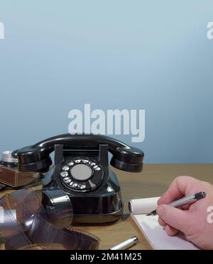 Die Hand des Mannes schreibt in ein Notizbuch mit einem Füllfederhalter. Altes Telefon, alte Kamera und Film auf einem Holztisch Stockfoto