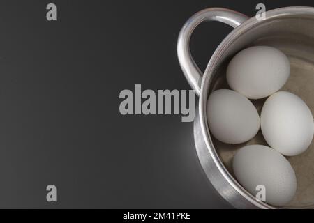 Metalltopf mit vier Eiern auf schwarzem Hintergrund, Draufsicht. Gekochte Hühnereier in einem Topf. Speicherplatz kopieren Stockfoto