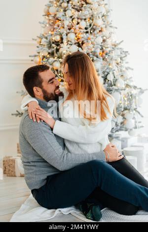 Ein attraktives Paar in der Nähe des Weihnachtsbaums und umarmt sich. Neujahrsatmosphäre. Zärtliche Umarmungen am Weihnachtsbaum zweier Menschen Stockfoto