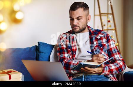 Ein Mann, der während der Weihnachtsfeiertage allein zu Hause arbeitet und sich Notizen in sein Notizbuch schreibt. Stockfoto