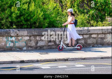 Eine junge Frau, die ein weißes Kleid trägt, fährt einen elektrischen Roller auf dem Bürgersteig. Stockfoto