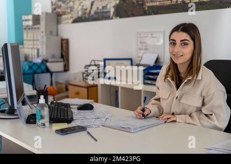 Eine junge Frau im Anzug, die auf einem Notizblock schreibt und telefoniert, während sie im Büro sitzt Stockfoto