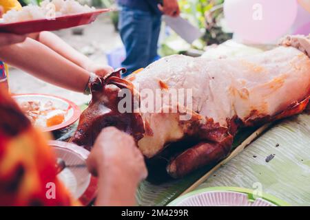 Metzger, der in Stücke schneidet, der allzeit beliebte Favorit, das beliebte köstliche, gebratene, knusprige ganze Schweinefleisch oder Lechon Baboy, lag auf Bananenblatt auf dem Tisch Stockfoto