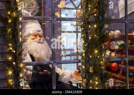 Der Weihnachtsmann im Retro-Stil sitzt hinter einem Glasfenster des Hauses, mit Weihnachtsdekoration einkaufen, Lichter. Hochwertiges Foto Stockfoto