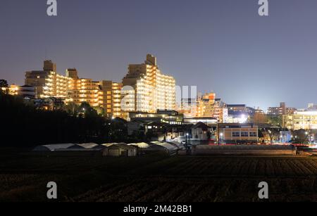 Gut beleuchtetes Apartmentgebäude über Wohngegend neben einer kleinen Farm in der Nacht Stockfoto