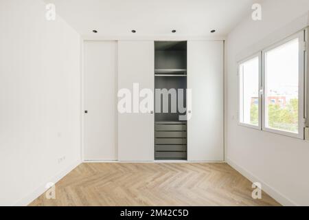 Zimmer mit eingebautem Kleiderschrank mit weißen Schiebetüren, weiß lackierten glatten Wänden, grauem Schrankinnenraum mit Schubladen und Eichenboden Stockfoto