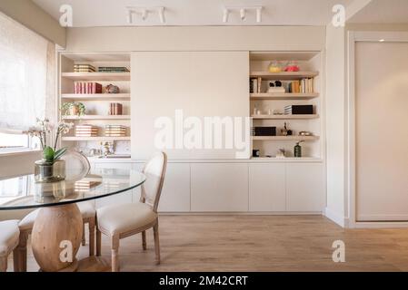 Ein Wohnzimmer mit einem hölzernen Bücherregal mit eingebauten Regalen, weißen Spots an der Decke und einem runden Glastisch Stockfoto