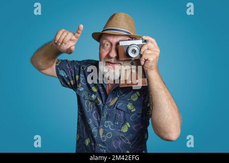 Sagen Sie, Cheese macht ein Foto. Glücklicher Seniorenmann, grauer Bart und Strohhut mit Retro-Kamera. Aufsteigendes Fingerporträt. Blauer Hintergrund. Stockfoto