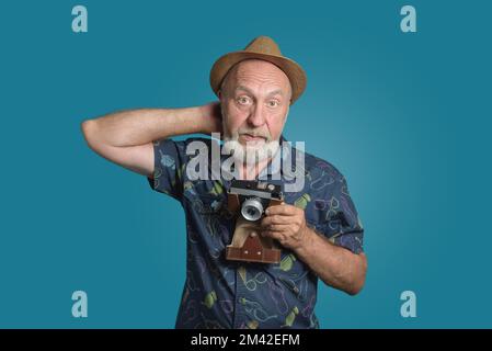 Seniorfotograf mit kratzendem Kopf einer Retro-Kamera. Hat seinen Strohhut zurückgeblasen. Frontales Porträt, dunkelblauer Hintergrund Stockfoto