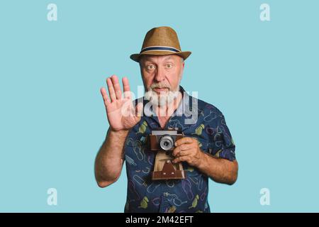 Posieren Sie. Ein alter Mann mit grauem Bart und Strohhut, der eine alte Kamera hält, hebt seine Hand hoch, um ein Foto zu machen. Blaues Hintergrundporträt Stockfoto