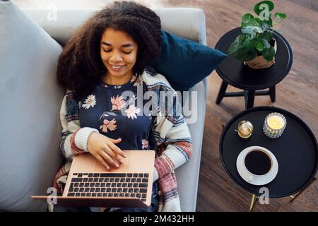 Aufgeregte, fröhliche junge schwarze Frau, die zu Hause einen Laptop auf dem Sofa benutzt, gute Nachrichten bekommt, Freude hat, Hausaufgaben macht oder online arbeitet, lacht, Stockfoto