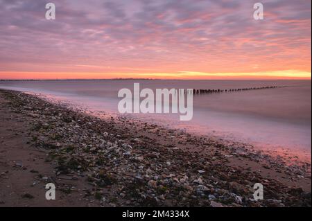 Sonnenaufgang über der Nordsee auf East Mersea Island in Essex. Wellenbrecher im Langzeit-Meer. Warmer, roter, orangefarbener und gelber Morgenhimmel. Lebendige Farben Stockfoto