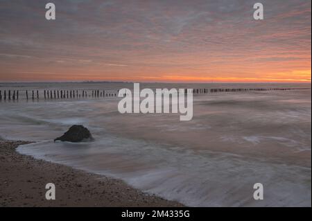Sonnenaufgang über der Nordsee auf East Mersea Island in Essex. Wellenbrecher im Langzeit-Meer. Warmer, roter, orangefarbener und gelber Morgenhimmel. Lebendige Farben Stockfoto