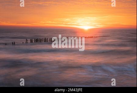 Sonnenaufgang über der Nordsee auf East Mersea Island in Essex. Wellenbrecher im Langzeit-Meer. Warmer, roter, orangefarbener und gelber Morgenhimmel. Lebendige Farben Stockfoto
