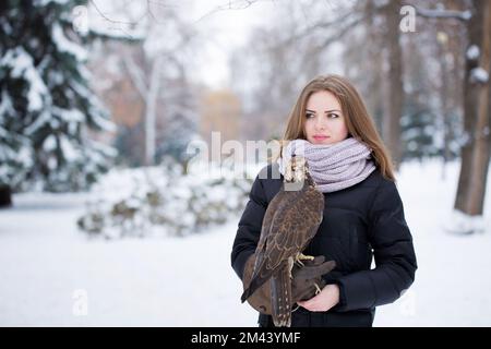 Ein Mädchen in einem schwarzen Mantel hält im Winter einen Falken an der Hand Stockfoto