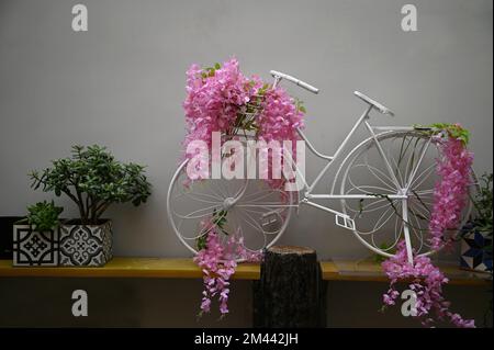 Klassisches weißes Fahrrad auf einem Holzregal mit pinkfarbenen Satinblumen an einer weiß getünchten Wand. Stockfoto