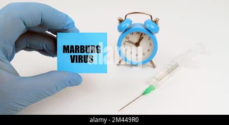 Medizinisches Konzept. Auf dem Tisch ist eine Spritze und ein Wecker, in seiner Hand ein Notizbuch mit der Inschrift - Marburg Virus Stockfoto