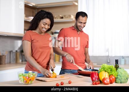 Lächelnder schwarzer Mann und Frau der Jahrtausendwende in roten T-Shirts schneidet Salat, bereitet Mittagessen zu, macht Hausarbeiten in der Küche Stockfoto