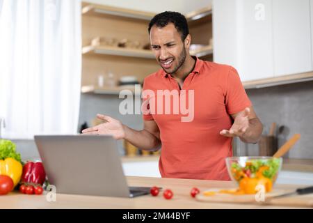Verärgerter junger schwarzer Mann in rotem T-Shirt, der Hände am Computer hat, hat Probleme mit dem Service Stockfoto