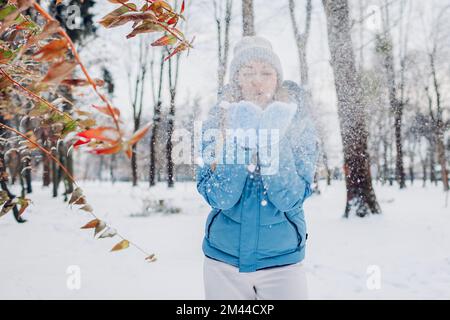 Glückliche junge Frau bläst Schnee aus den Händen, um Spaß im Winterwald zu haben. Mädchen mit blauem Mantel und Fäustlingen. Spielen im Freien bei kaltem Wetter Stockfoto