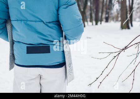 Rückansicht einer Frau, die im verschneiten Winterpark in einer blauen Daunenjacke mit reflektierendem Streifen und Klebeband läuft. Warme Kleidung für kaltes Wetter Stockfoto