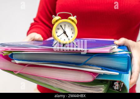 Büroangestellter, der einen Stapel von Dokumenten mit Wecker, Konzept, Termin, Arbeit unter Druck, Fristeneinhaltung, Überstunden, Stress bei der Arbeit, Stockfoto